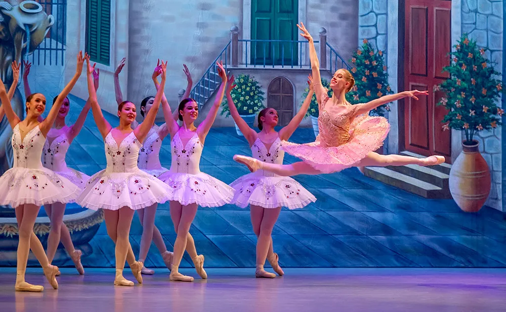 Ballet performance featuring a female soloist performing a saut de chat with corps de ballet on stage